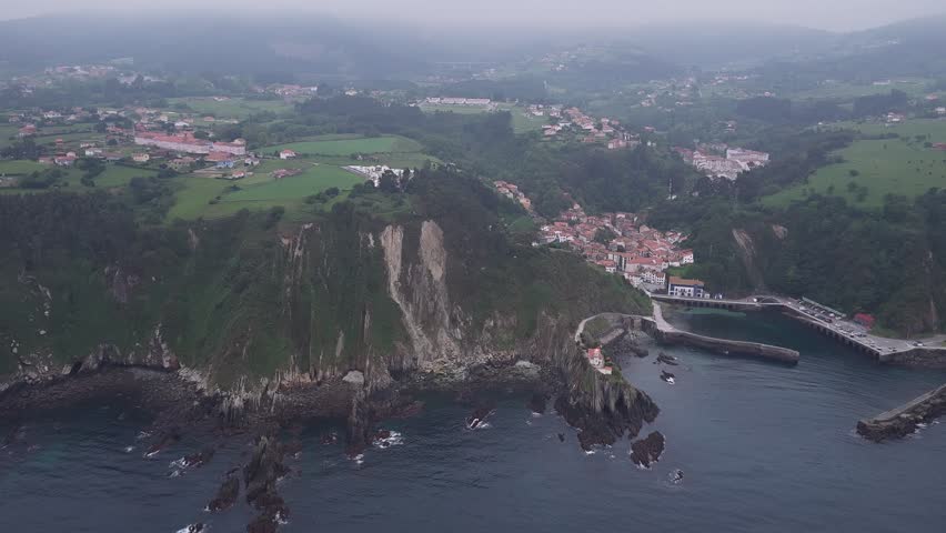Aerial view of the colorful houses of Cudillero village tucked into a lush green valley along the rugged rocky coastline under a misty sky Cudillero, Asturias, Spain.