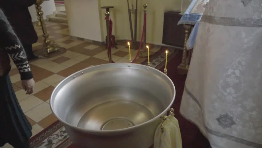 View of large metal baptismal font filled with water, positioned on red carpet in church interior with tiled floor. Lit candles burn near font, suggesting upcoming religious ceremony, people stand nea