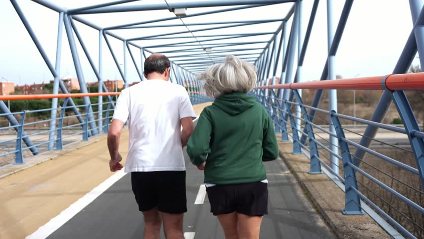 Active senior couple enjoying a healthy lifestyle while jogging together across a modern pedestrian bridge. Rear view of two people in love