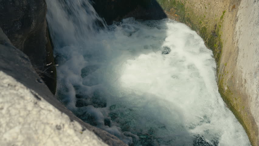 White water churns below a narrow waterfall, filling a rock basin with foam, motion, and bright natural energy inside a smooth stone channel.