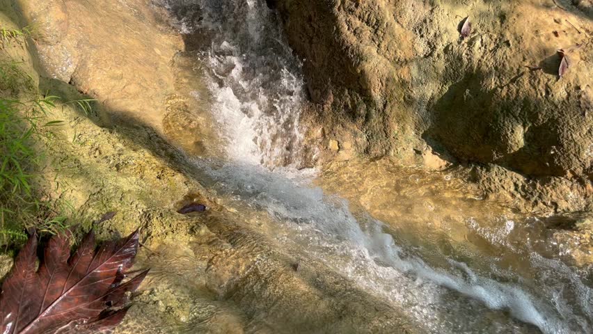 Close-up diagonal shot of clear water rushing over textured limestone rocks in a forest with a large dry leaf in the foreground.