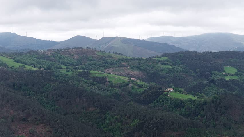Aerial view of lush green hills, dense forests and wind turbines on the horizon under an overcast sky in Asturias, Asturias, Spain.