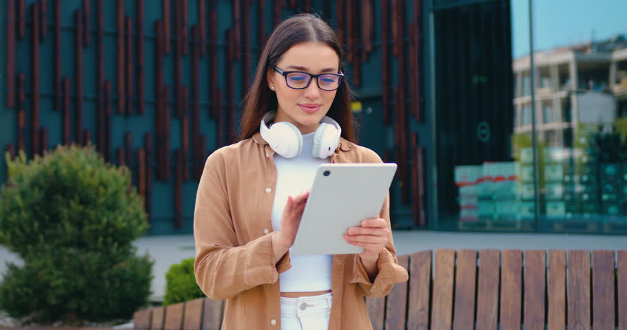 Portrait of a young businesswoman wearing glasses and casual attire, outside a modern office building, scrolling through a tablet with a happy expression and reading good news