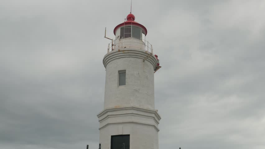 Handheld close up view of a white maritime lighthouse with a red dome in Vladivostok. The navigation tower stands against a gloomy overcast sky.