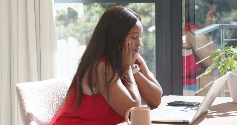 African American woman sitting at table beside glass door, cupping face, reading laptop for work. Focus, contemplation, homeoffice, daylight, window, nature, minimalist