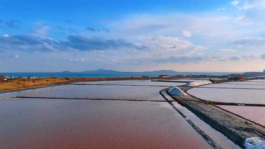 Salt ponds mirror sky and water. Salt ponds stretch across the land under a blue sky with clouds during late afternoon. Water reflects nearby hills.