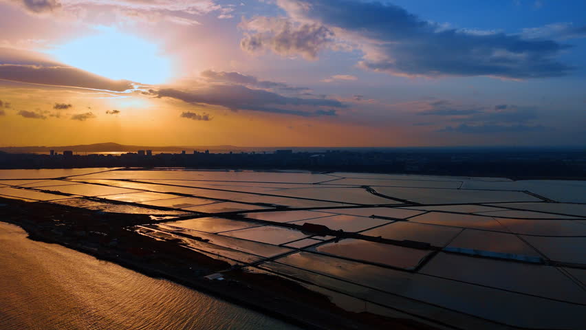 Vibrant sunset over rectangular salt pans near city skyline. Golden hour aerial shot of flooded salt evaporation ponds reflecting sunlight with urban buildings on the horizon.