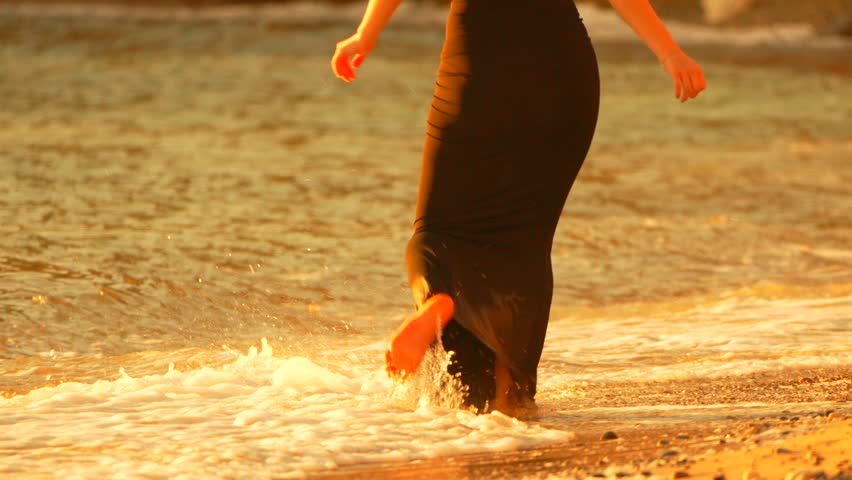 Woman beach sunset walking along the shoreline in a long black dress during the golden hour as waves wash over the sand