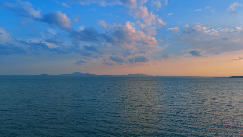 Sunset over water and clouds in the sky. Waves meet the horizon as sunlight fades and clouds float above the water. The day turns to evening in this calm view.