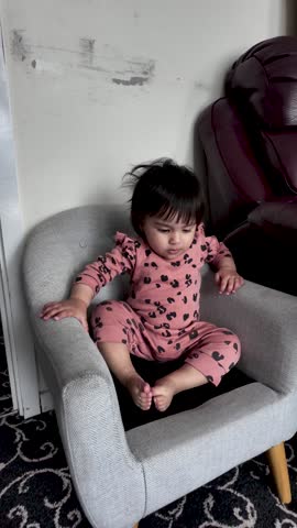 A cute South Asian baby girl is captured sitting on a small grey chair indoors, showcasing her early childhood development and innocent curiosity. She is dressed in pink pajamas with a leopard print.