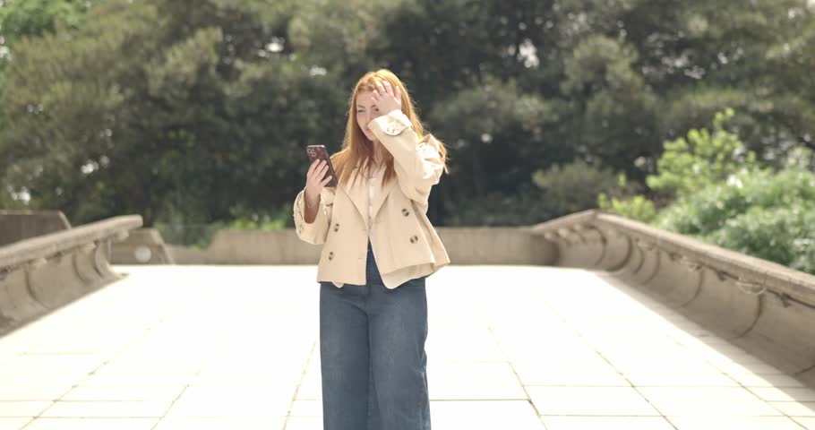 Cheerful beautiful woman with long red hair walking on a bridge in the park and using a smartphone
