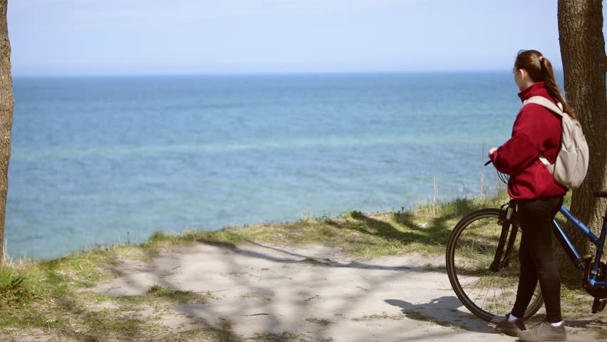 Adult female cyclist wearing red jacket and white backpack stands on coastal dirt path next to black bike, surrounded by green grass and two tree trunks with calm water horizon background