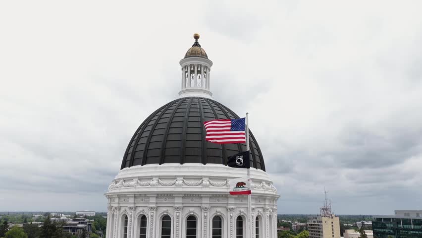 California State Capitol dome with waving American flag in Sacramento, United States, with neoclassical architecture, cloudy sky and patriotic atmosphere in government landmark. Aerial orbiting shot.