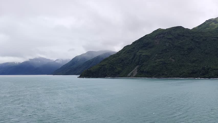 Alaska, USA - View of the Hubbart Glacier and coastline. The scene captures a mountain range partially covered in green vegetation along the calm ocean water.