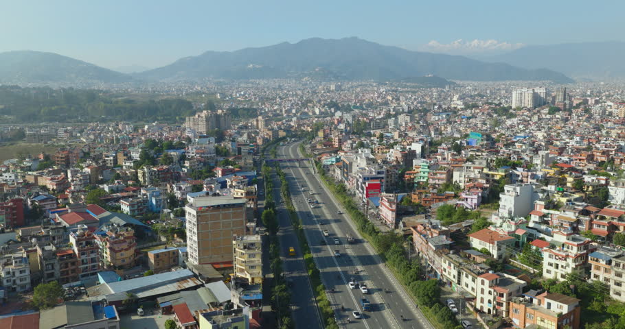 Aerial view of the unmanaged city of Kathmandu, Nepal, with Ringroad highway cutting through buildings and clear mountains in the background under the blue sky.