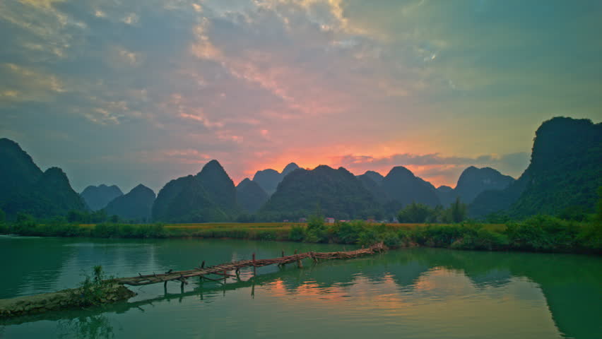  A serene sunset scene with golden light breaking through the clouds, casting reflections on a calm lake surrounded by lush green mountains. A rustic wooden bridge adds charm to the peaceful natural landscape.
