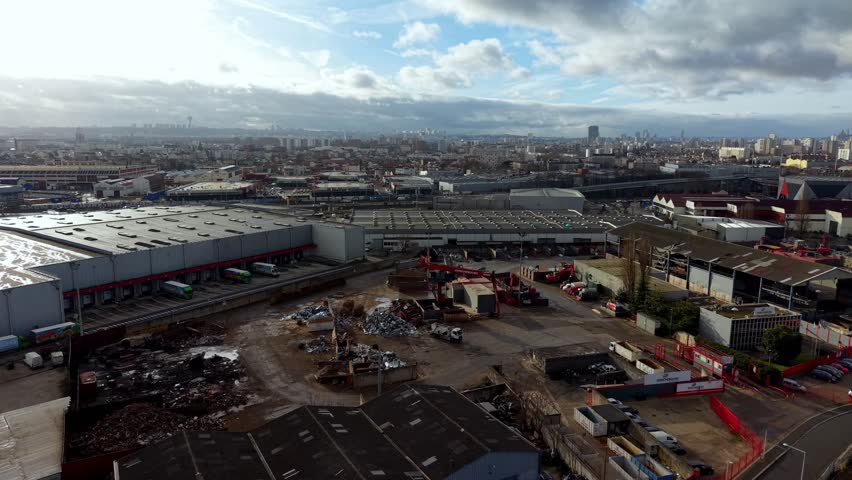 Logistics center and scrapyard in an industrial suburb with Paris city skyline on horizon, France. Aerial forward