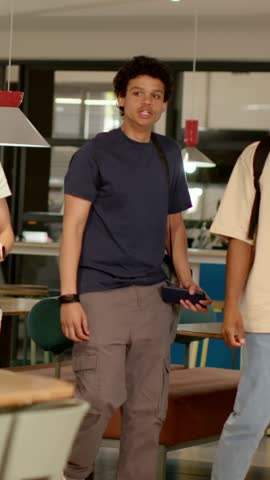 Vertical video: Walking in, man in navy tee with phone and bag shaking hands with friend in lounge. Handshake, greeting, casual, satchel, crossbody, device, vertical video