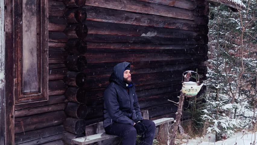 Man sitting on a wooden bench in front of a log cabin during snowfall in winter forest.