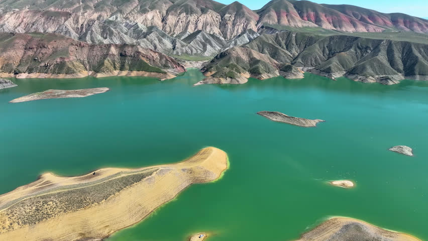 Aerial view of a turquoise lake surrounded by colorful mountains. Rocky shores and small islands dot the water