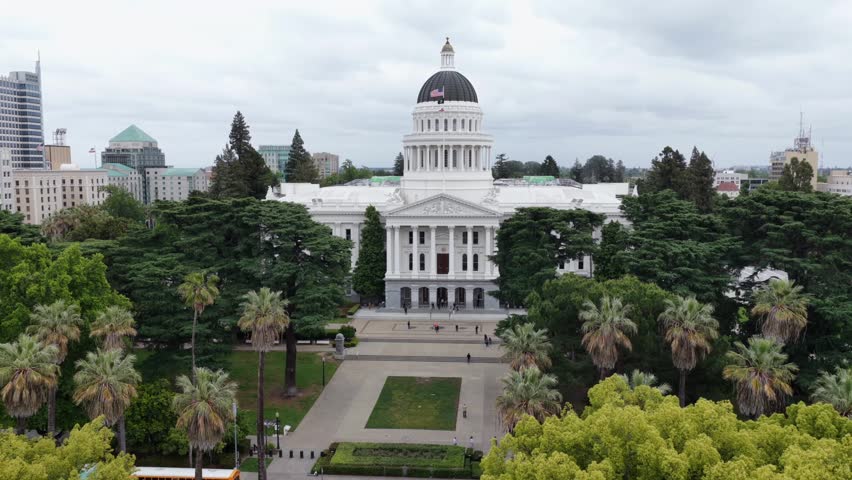 Aerial drone view of California State Capitol building in Sacramento, United States, with iconic white dome, American flag and surrounding palm trees in historic government district under cloudy sky.