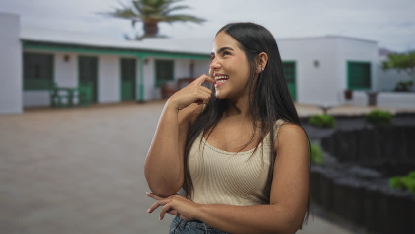 Young woman smiling and pressing finger to her teeth, standing in a sunlit courtyard in front of a white building with green doors and a palm tree; playful confidence.