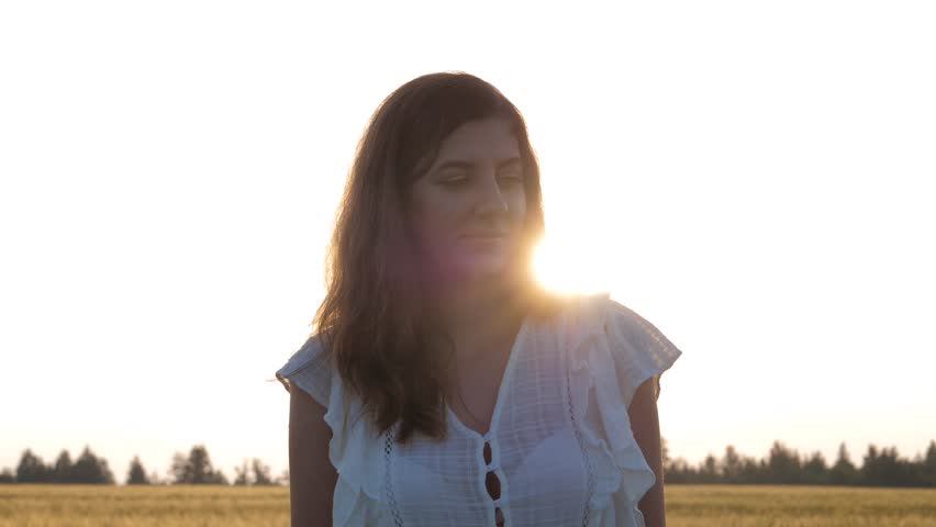 Pretty young woman with long hair in white dress standing in wheat field at summer sunset. Woman stretching hands up, smiling and looking at camera. Female enjoying freedom and summer nature