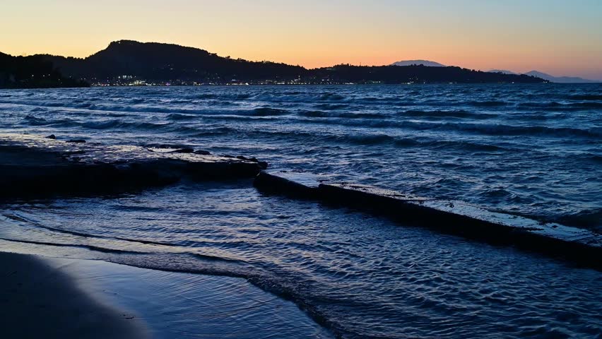 Ground level view of sea waves washing on the shore at dusk. Scenic twilight landscape with orange horizon, dark blue water, and city lights twinkling in the distance. Calm evening atmosphere.