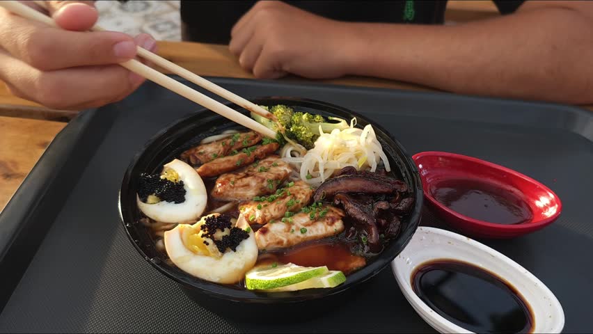 A detailed close-up shot of a rich ramen bowl served on a black tray. The bowl contains savory broth, thick noodles, grilled chicken strips, soft-boiled eggs topped with black caviar (tobiko), mushrooms, and fresh greens. A person is shown picking up noodles with wooden chopsticks. Two small side dishes with soy sauce and spicy dip are visible in the background.