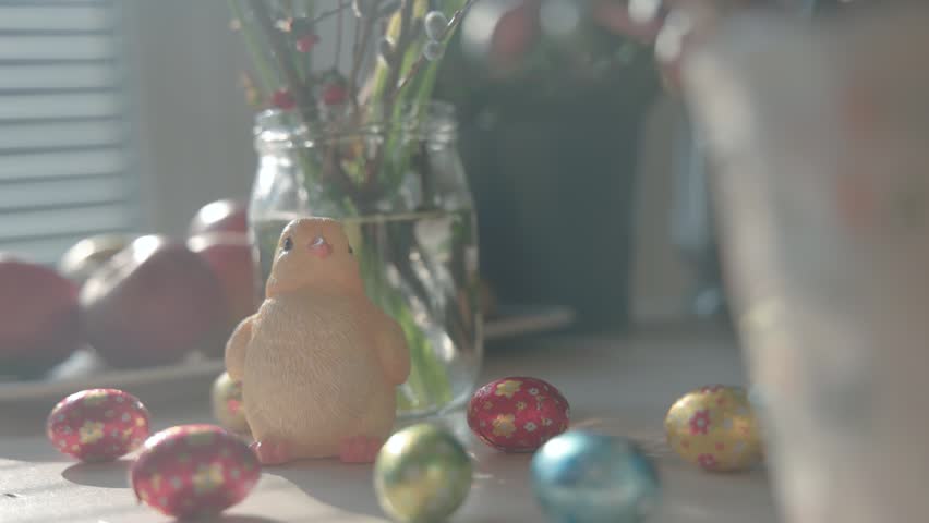 Cute yellow chick figurine on a sunlit table surrounded by colorful Easter eggs, with soft bokeh and shallow depth of field creating a warm, festive spring atmosphere indoors.