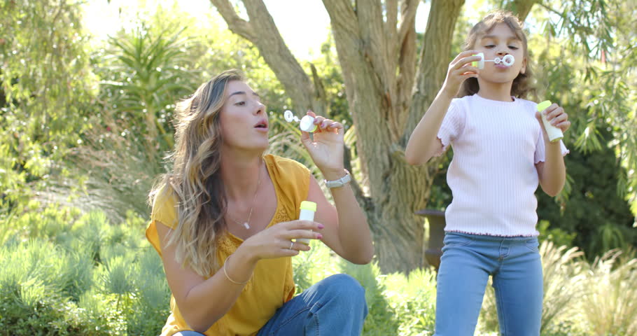 Mother and daughter playing as woman starting blowing bubbles, dipping wands and bottles in garden. Backyard, outdoors, sunny, family, girl, grass, tree