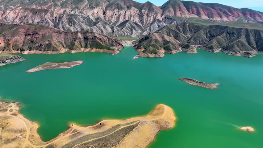 Aerial view of vibrant green lake surrounded by colorful rocky mountains. Sandy shores curve along calm water surface. Mountains display red, brown, and gray hues
