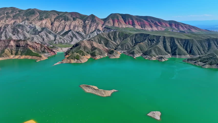 Aerial view of vibrant green lake surrounded by colorful mountain ranges. Mountains display red, orange, and gray tones. Clear blue sky above calm water surface