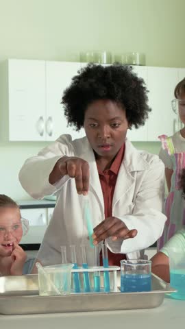 Instructor picking up blue foam tube, inserting into test tubes, scooping powder, teaching science. Classroom, lab, goggles, mentor, pupil, stem, vertical video
