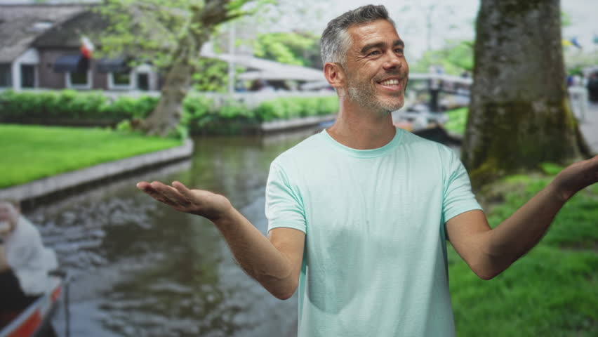 Man middle age grey hair smiling with outstretched palms beside a canal on a street wearing light teal t shirt; optimism.