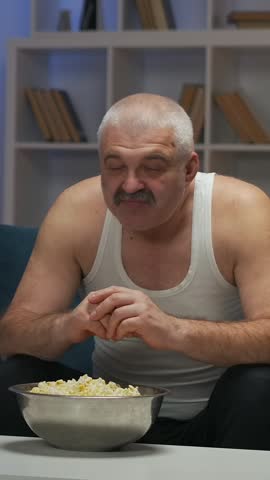 Older man in white tank top watching television at home, reacting emotionally as he eats popcorn from a metal bowl while seated on a living room couch in the evening