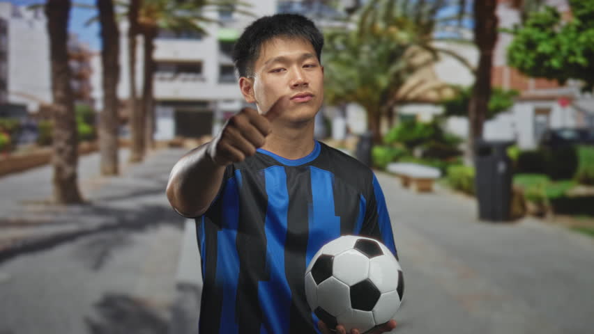 Young chinese man in blue soccer uniform holding a soccer ball and giving thumbs down on a sunny street; disapproval rejection.