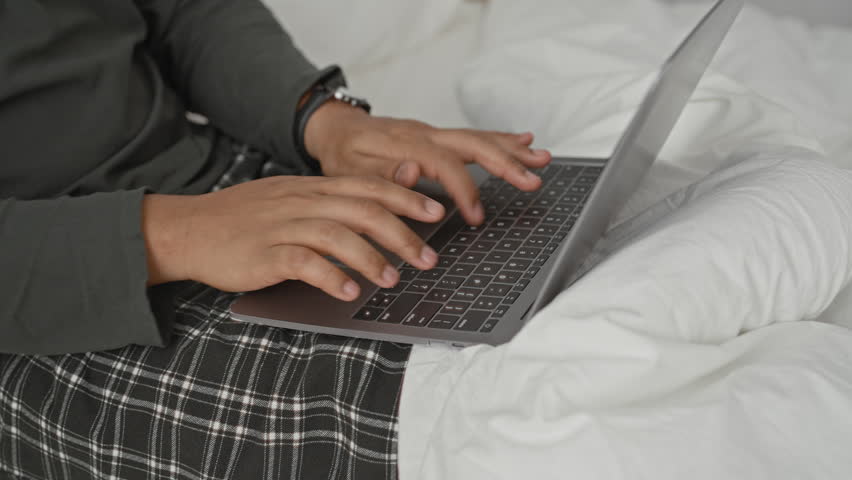 Man typing on laptop with hands on keyboard in building bedroom by a bed and nightstand; concentration remote work.