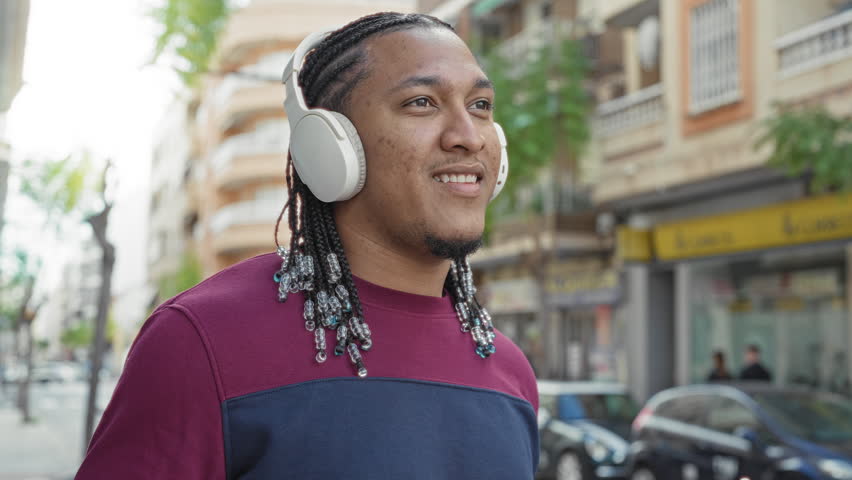 Man with headphones touching earcup and raising hands, smiling with beaded braids and maroon sweatshirt on a busy city street; joyful relaxation groove.
