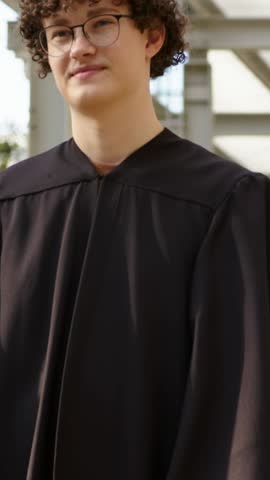 Vertical video: Smiling grad in black gown raising cap after name call at campus with gold tassel. Man, mortarboard, glasses, curlyhair, university, portrait, vertical video