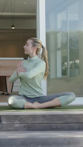 Vertical video: Breathing woman in green top and leggings twisting on porch with yoga mat for calm. Adult, wellness, minimal, fitness, steps, outdoors, vertical video