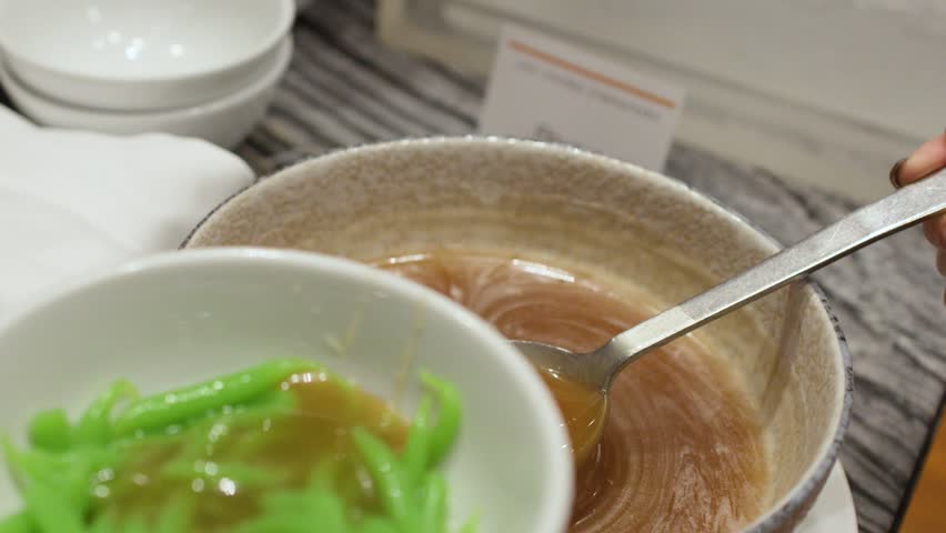 Close-up shot of brown palm sugar syrup poured onto green pandan noodles in white bowl