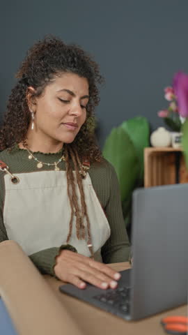 Woman florist in studio wearing apron smiles with hand on laptop and tulips on counter near brown paper roll; joyful entrepreneurship.