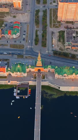 Vertical video. Yoshkar-Ola, Russia. The Bruges Embankment stretches along the Malaya Kokshaga River, its Flemish-style buildings with stepped gables, bright facades and decorative turrets forming a distinctive waterfront ensemble. At the center stands a replica of the Spasskaya Tower of the Moscow Kremlin, acting as a gateway between the two riverbanks. The embankment was developed between 2010 and 2014 and has since become the city