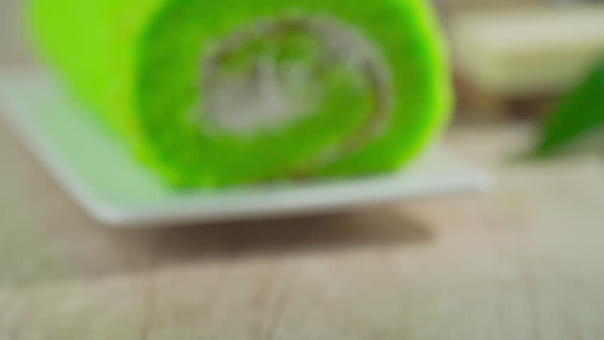 Pandan cake roll rests on white plate. Freshly sliced, revealing creamy white filling inside. Surrounded by eggs, butter, sugar, and pandan leaves. Natural wood surface gives warm, rustic feel.