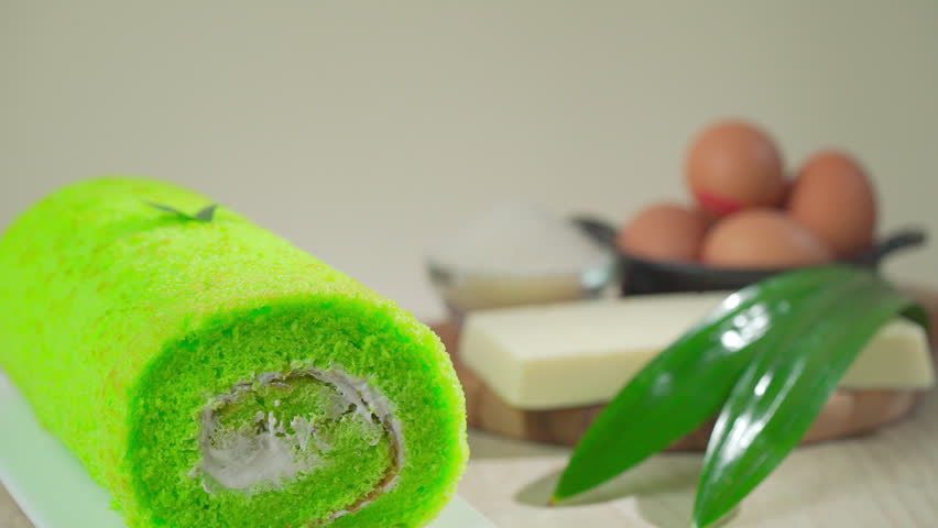Pandan cake roll rests on white plate. Freshly sliced, revealing creamy white filling inside. Surrounded by eggs, butter, sugar, and pandan leaves. Natural wood surface gives warm, rustic feel.