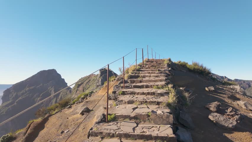 Tracking shot follows a stone mountain ridge path with metal posts, revealing a sea of clouds and rugged peaks at Pico do Arieiro on Madeira island, Portugal.