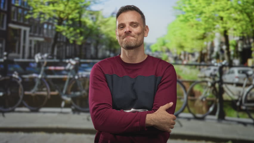 Man with hand on chin and crossed arms smiling on a street by a canal with bicycles and bridge; thoughtful reflection.