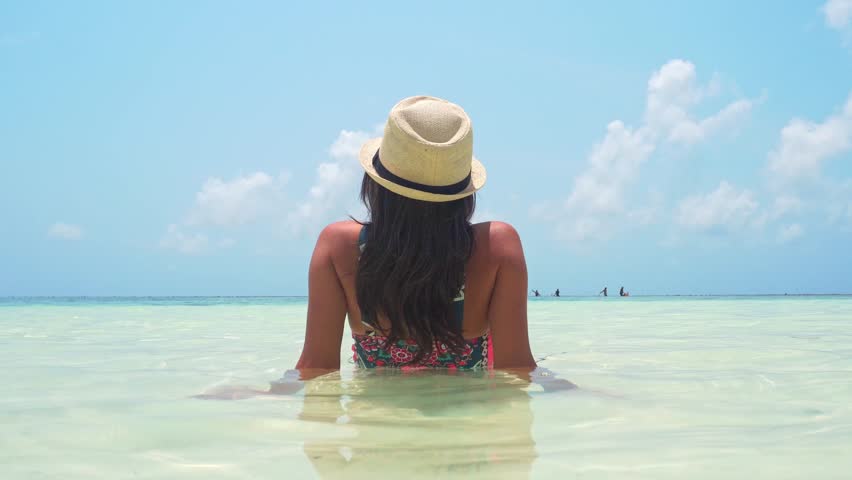 Pretty caucasian woman traveler tourist wear straw hat sits in crystal-clear, shallow turquoise water on a tropical beach. High-angle shot capturing pure serenity and vacation relaxation in paradise.