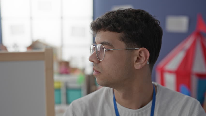 Young man with glasses and blue lanyard looking sideways near a whiteboard and playhouse in a preschool classroom; calm focus patience care.